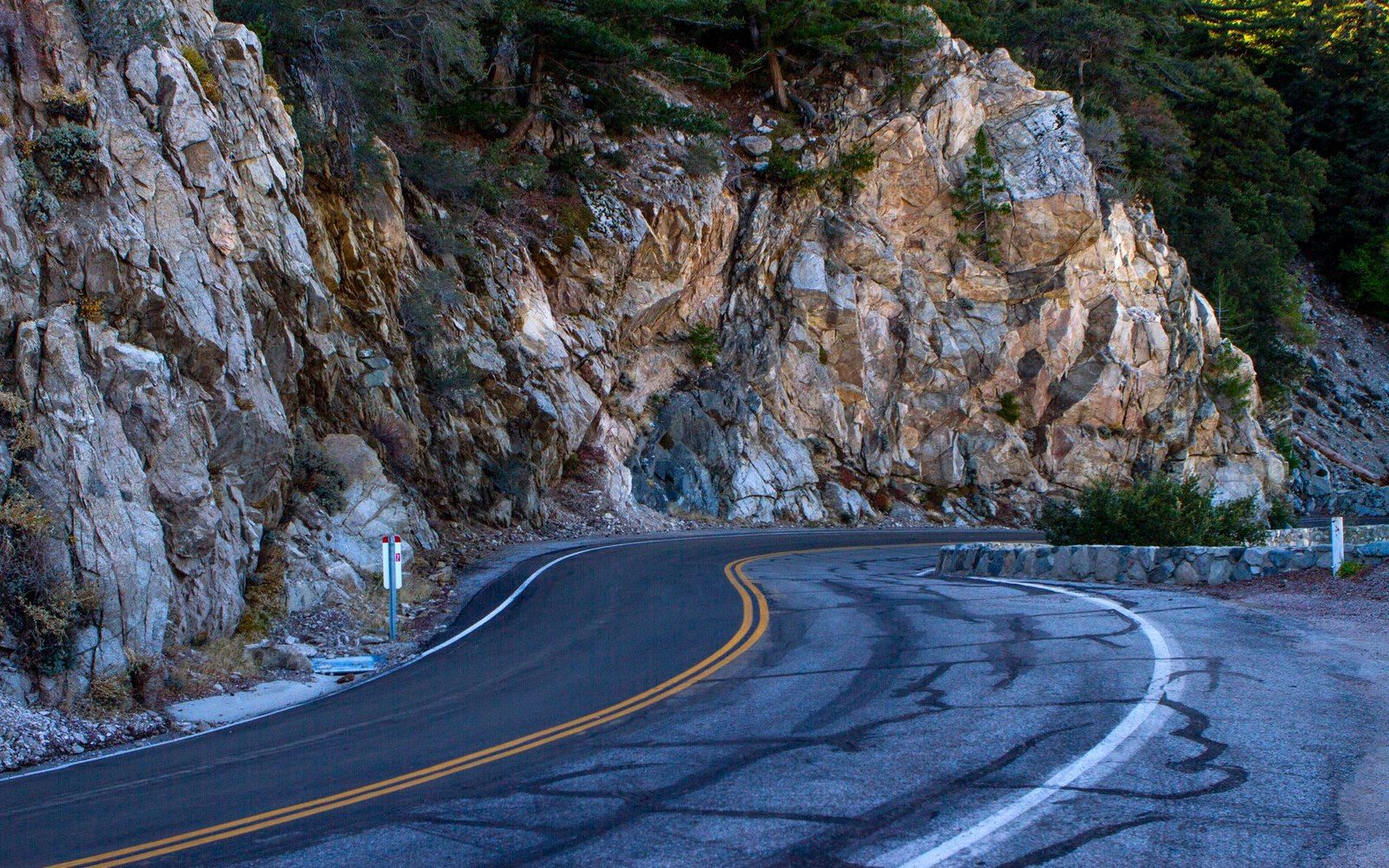 A winding mountain road curves past a jagged rock cliff carved into the hillside