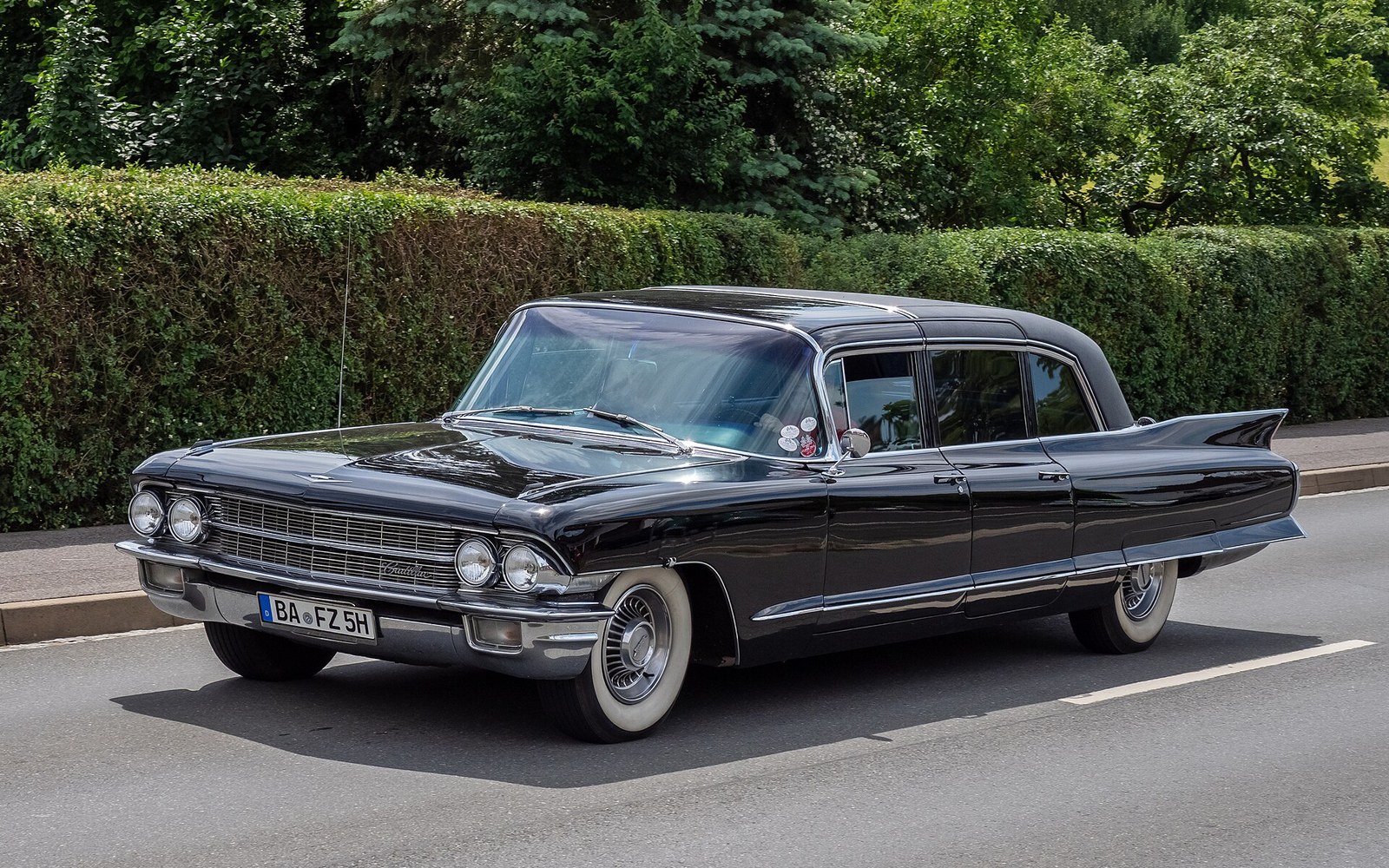 A vintage black Cadillac Fleetwood 75 limousine parked along a hedge-lined street
