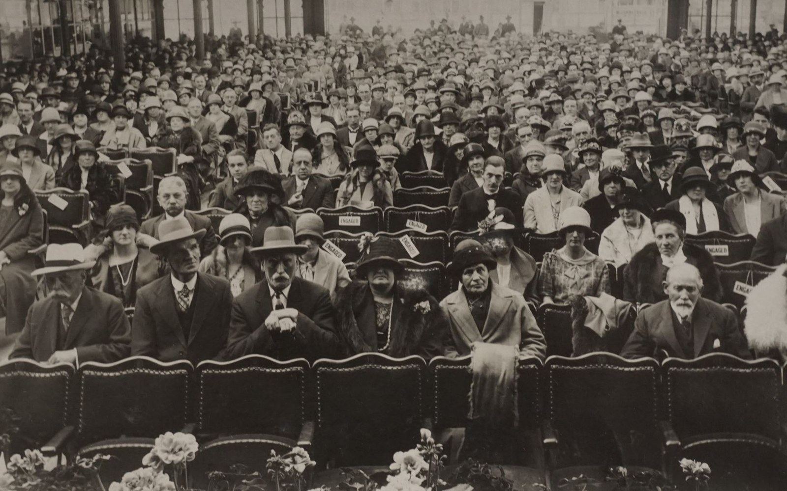 Rows of hatted men and women seated in an outdoor auditorium, most wearing formal early 20th-century attire