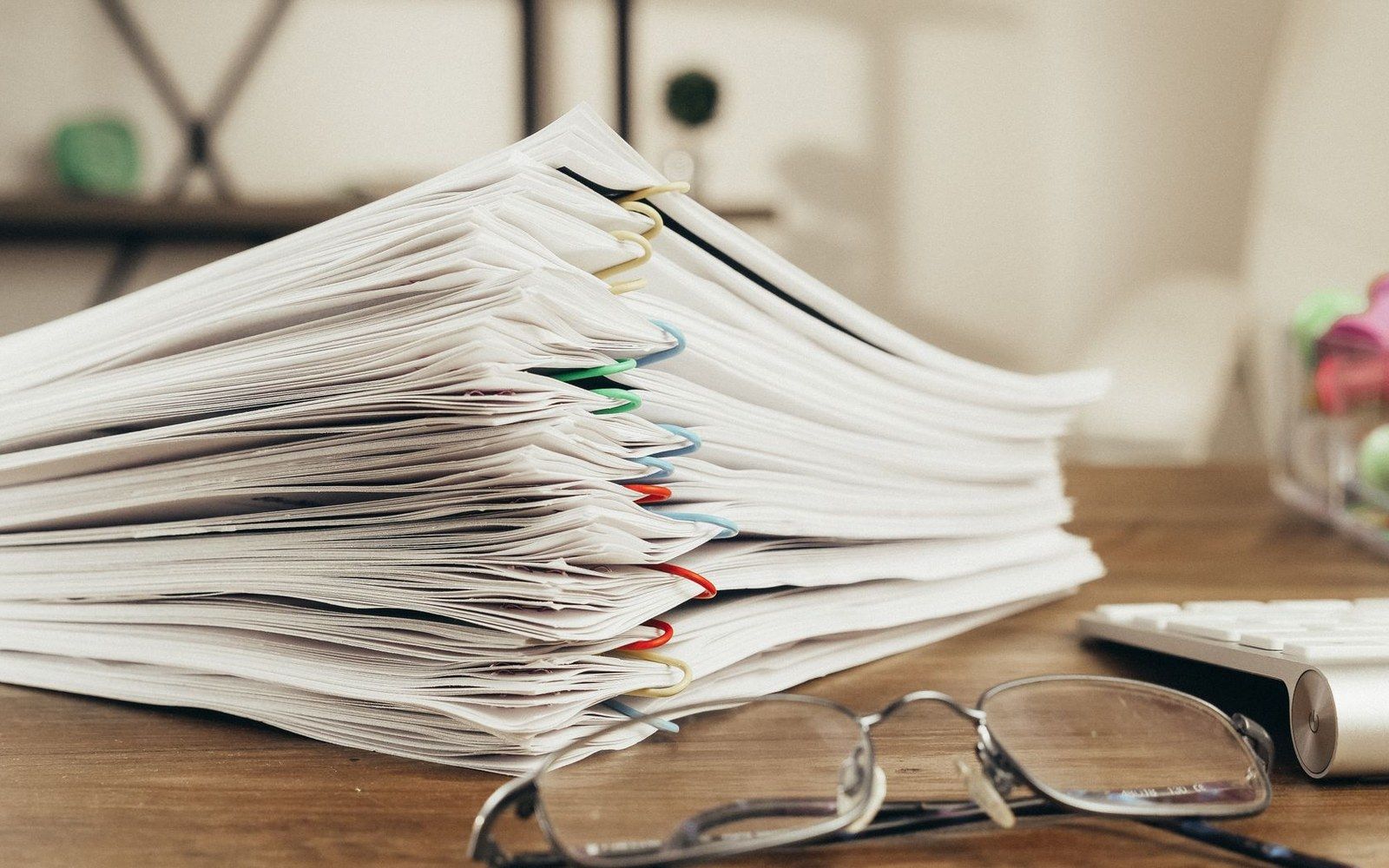 Stacked paper documents bound with colorful paper clips beside eyeglasses on a wooden desk