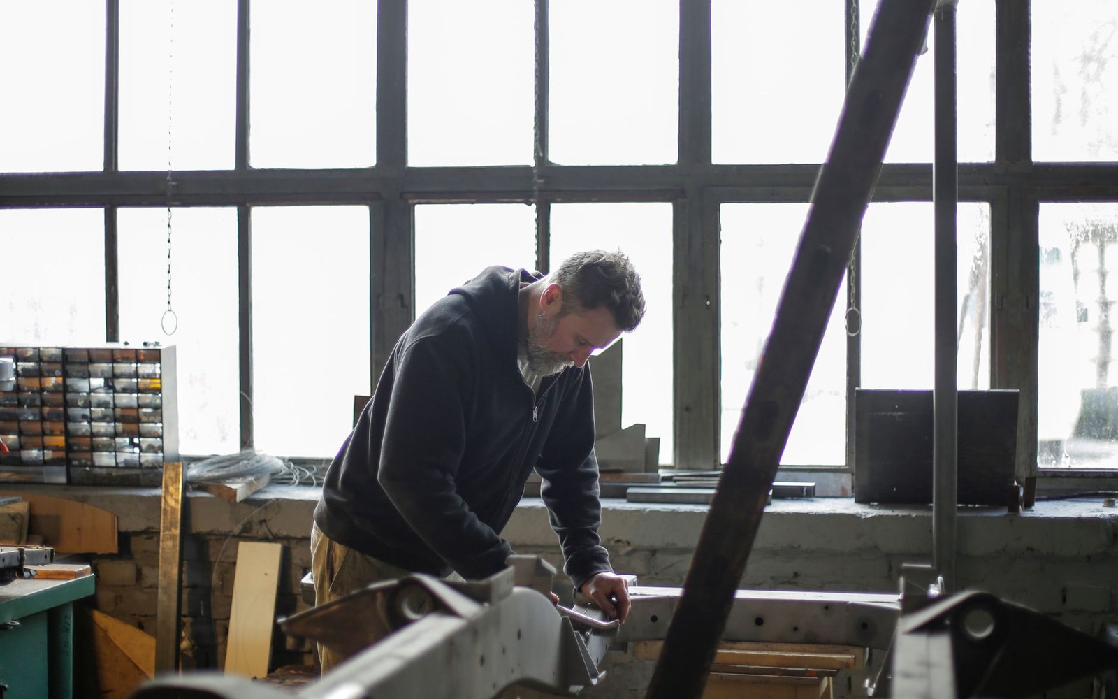 A man works on metal machinery in a workshop lit by large industrial windows
