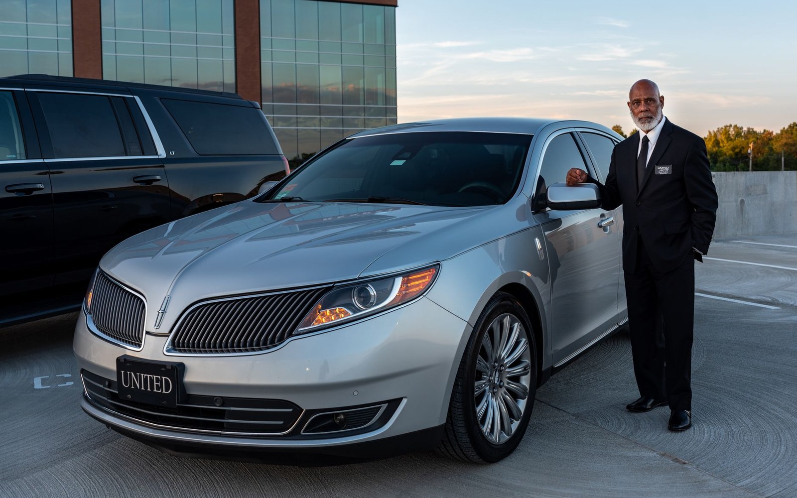 A chauffeur in a suit stands beside a silver sedan in a parking garage